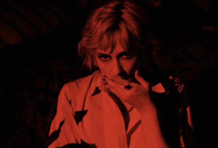 Young blonde woman drenched in red lighting, She is covering her mouth, her eyes suken.